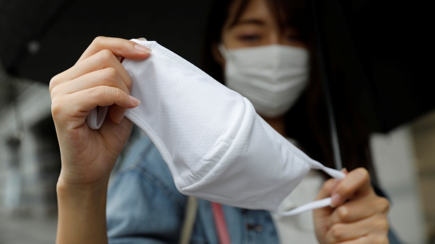 A woman wearing a white face mask holds a second mask up closer to the camera.