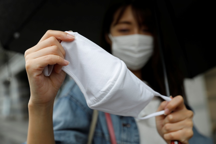 A woman wearing a white face mask holds a second mask up closer to the camera.