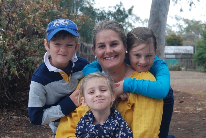 a woman sits on a blanket outdoors, with her three children climbing on her.