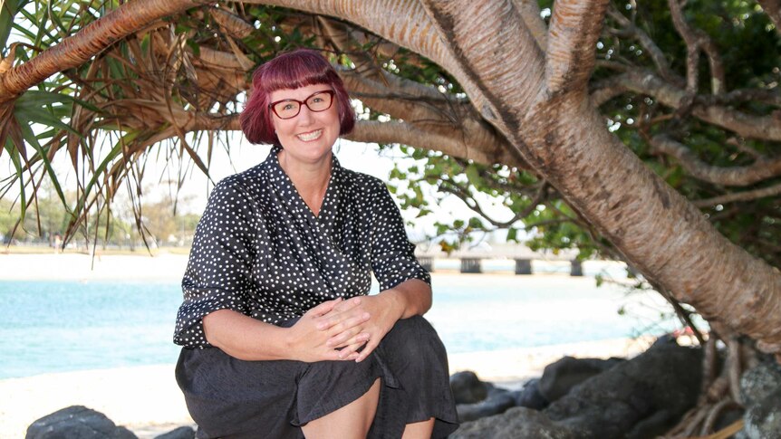 A woman with short red hair, wearing a black and white spotted blouse and glasses, sitting under a pandanus tree near the water.