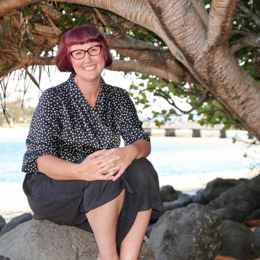 A woman with short red hair, wearing a black and white spotted blouse and glasses, sitting under a pandanus tree near the water.