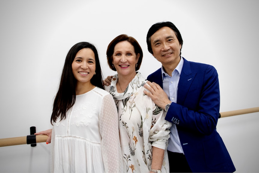 A man and a woman stand at a ballet bar with white wall next to their adult daughter. All are smiling