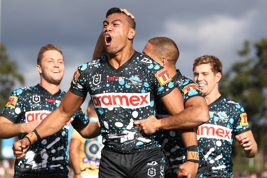 Ronaldo Mulitalo shouts after scoring a try for the Sharks. His teammates are hugging him from behind.