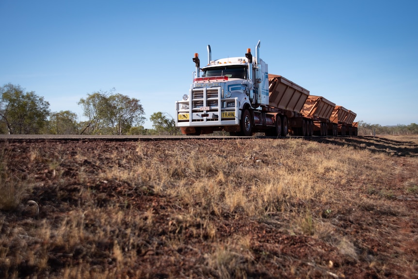 A road train with four trailers drives along a one-lane road with a steep verge.