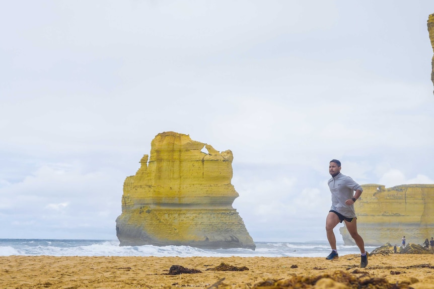 A man runs on beach in front of a large rock formation rising out of the sea.