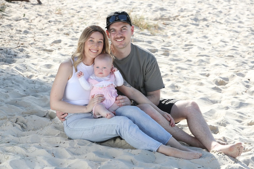 Sarah Shaddick with baby daughter Halle and partner, Luke Hill, sitting on the beach