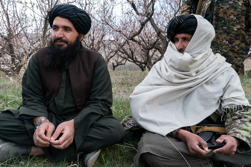 A man in black robes and another in white sit cross legged in an orchard