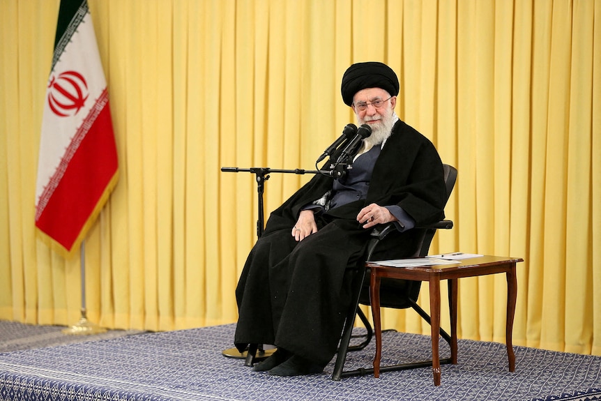 Man dressed in black speaking into microphone while seated on stage, with Iranian flag in background.