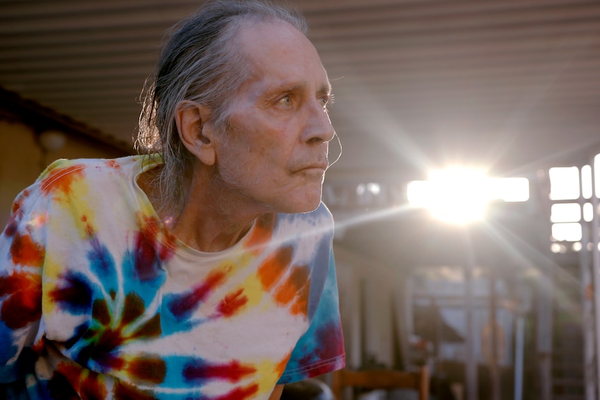 Gary Young wears a tie-dye shirt and looks out over a porch as sunlight pours in from behind