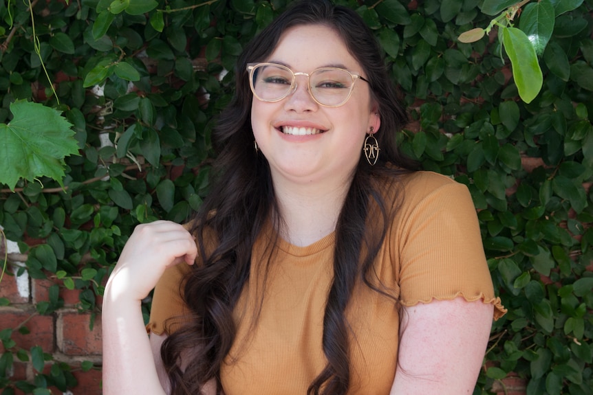 A woman in an orange top stands and smiles with a leafy green wall behind.