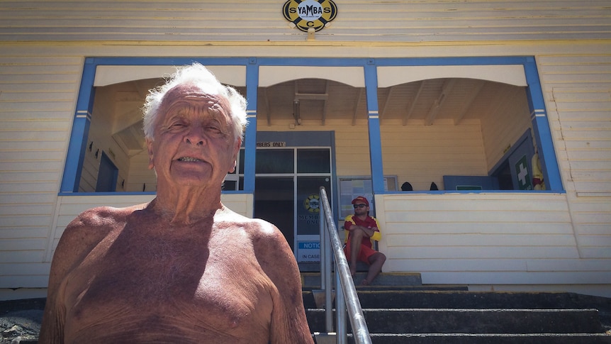 Man with no shirt on stands in front of old wooden building