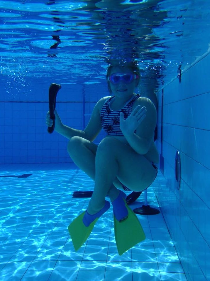 An underwater shot of a girl wearing flippers and goggles and holding a mini hockey stick, waving at the camera.
