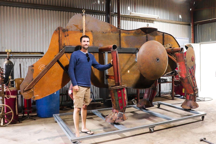 A man in shorts and a long-sleeved top stands next to a large, unfinished metal sculpture.