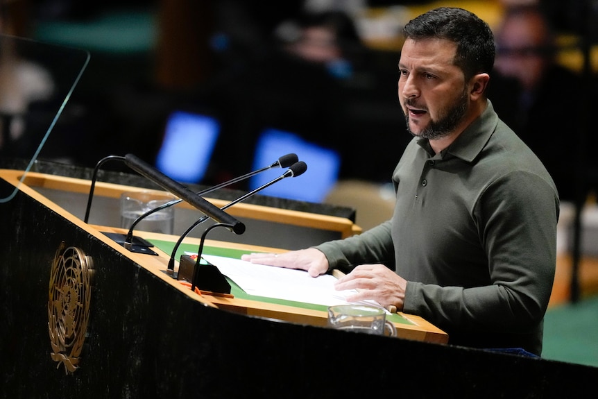 Volodymyr Zelenskyy, wearing an olive polo shirt, speaks into two microphones at a UN podium.
