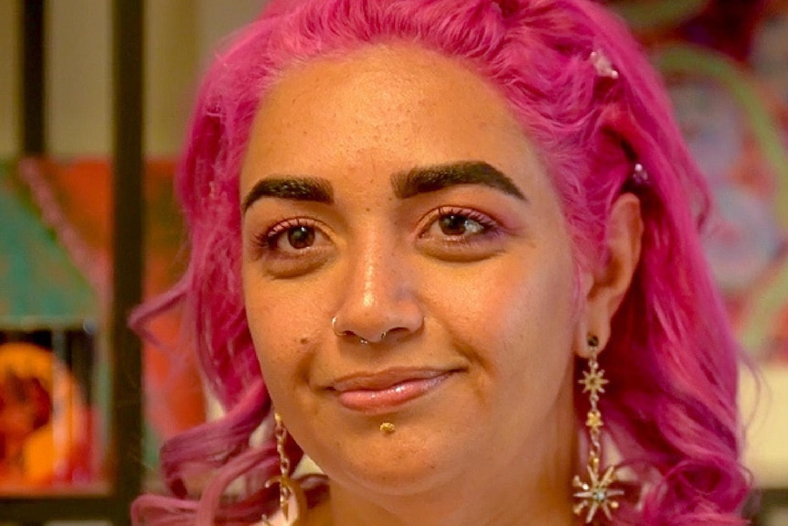 A woman with pink hair and sparkly, dangling earrings looks at camera