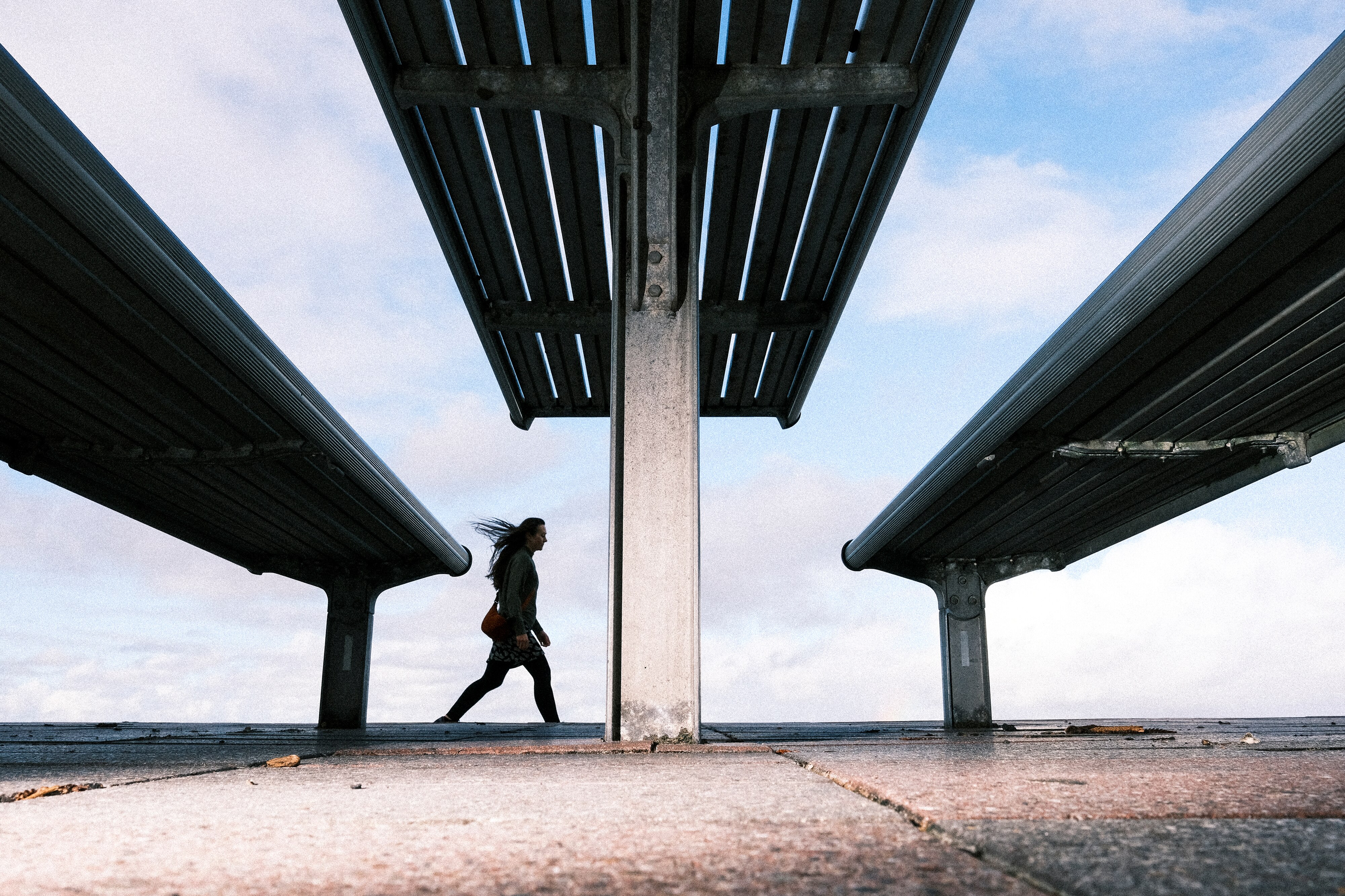 A low wide shot of metal benches with a metal table dwarfing a silhouetted person walking along a path near a beach.