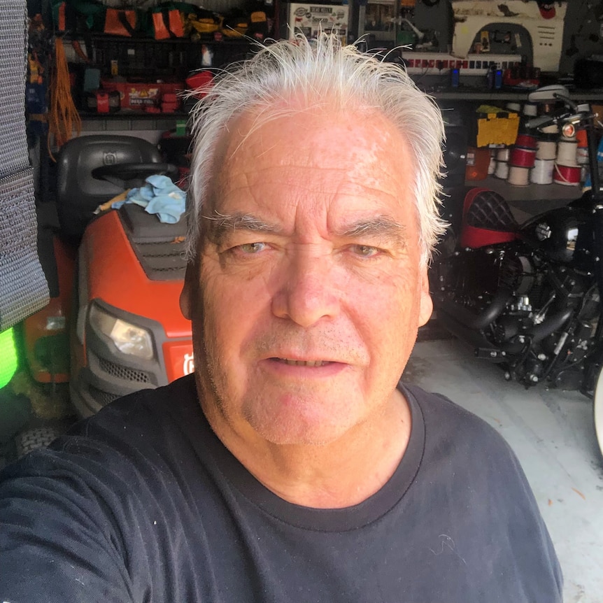 A man stands in front of an open garage with a motorbike behind him.