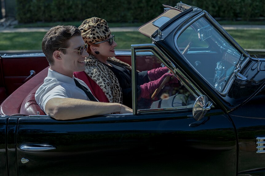 A man with sunglasses sits smiling next to older female driver wearing leopard print attire in vintage convertible on sunny day.