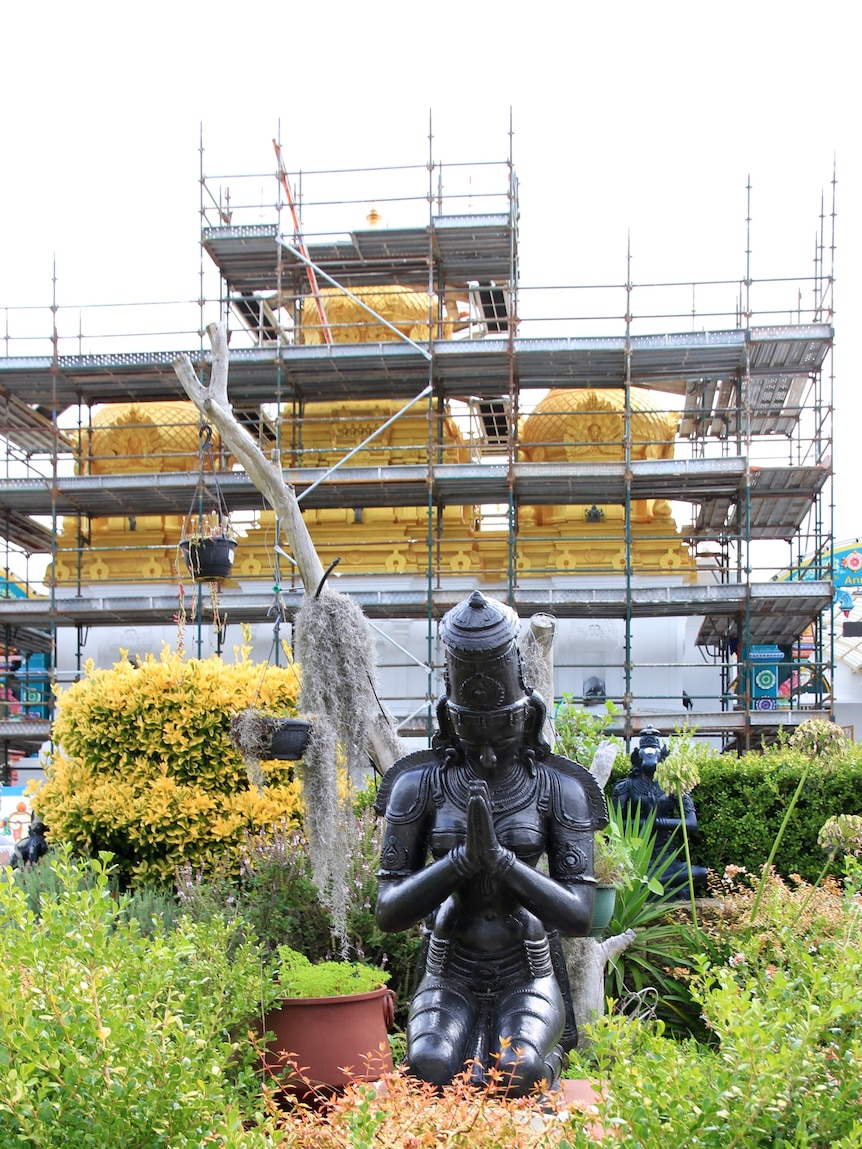 Scaffolding covers a gold and white temple, and in the foregound a statue is praying