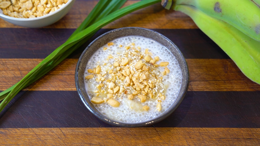 A bowl of banana sago pudding topped with crushed peanut, with Pandan leaf and green bananas alongside the dessert.