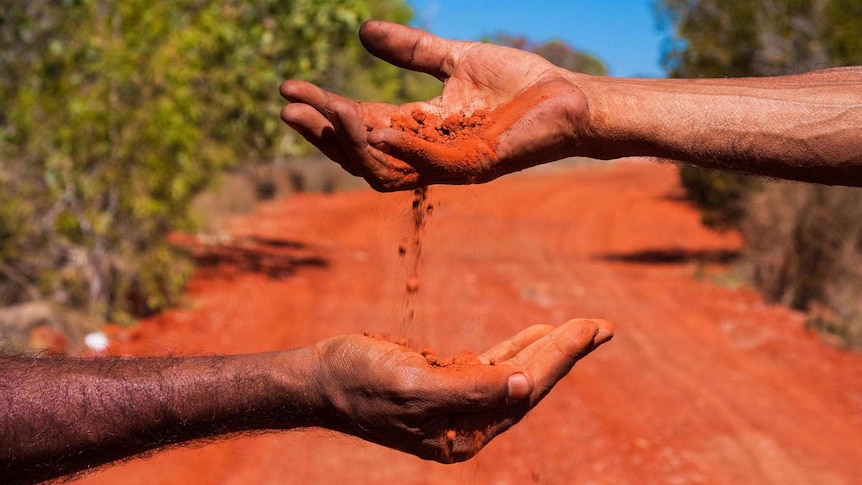 Red dirt falling from an unseen person's hand into another unseen person's hand. A red-dirt road is in the background.