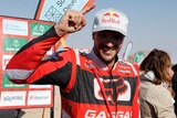 A motorcycle rider stands in the sun wearing a cap and smiling with one finger pointing up in celebration after a race.