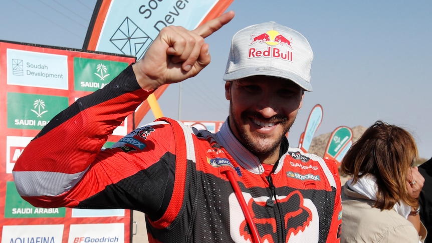 A motorcycle rider stands in the sun wearing a cap and smiling with one finger pointing up in celebration after a race.