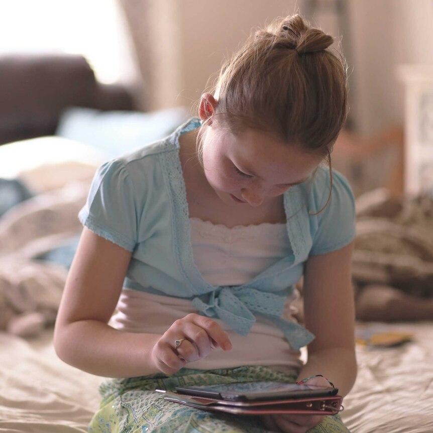 A young girl sits on a bed looking down at a tablet. There are pillows on the bed behind her and a brown couch in the background