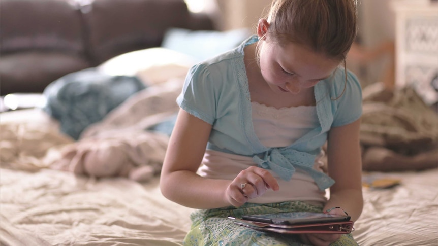 A young girl sits on a bed looking down at a tablet. There are pillows on the bed behind her and a brown couch in the background