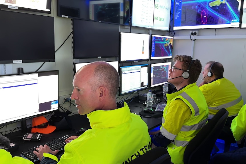Three men in hi-vis jackets sitting at computer terminals in front of a wall of screens, two on the right with headphones.