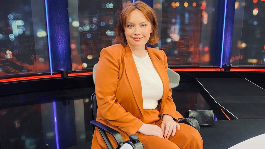 A smiling young woman in an orange pantsuit, sits on a wheelchair, behind her a wall of glass shows blurred city lights.
