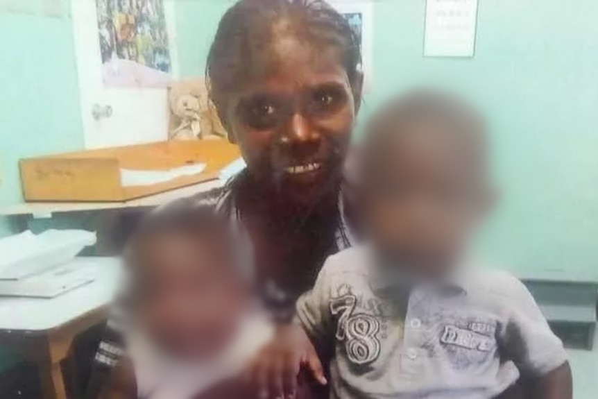 A young Indigenous woman sits with two small children whose faces are blurred