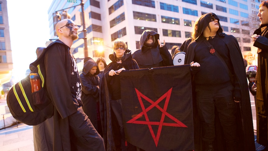 Satanists in street wearing black, holding black flag with pentagram.