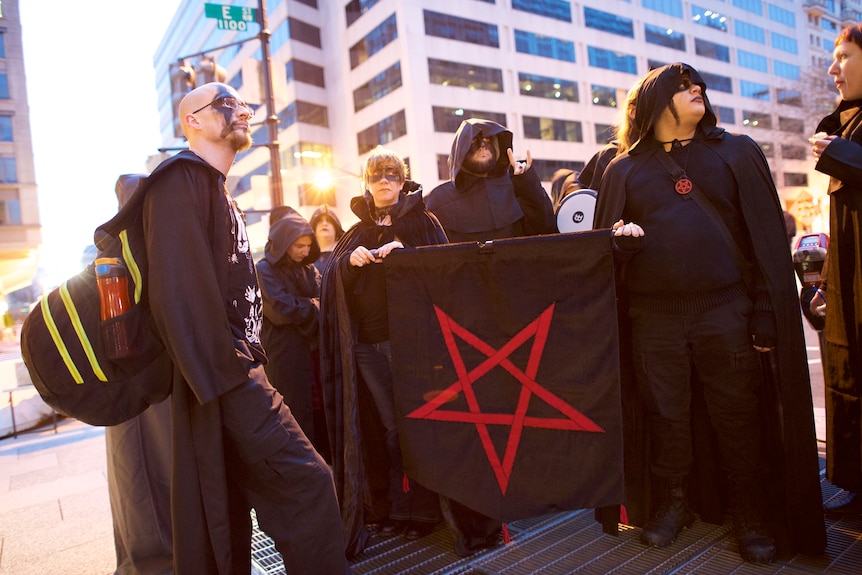 Satanists in street wearing black, holding black flag with pentagram.