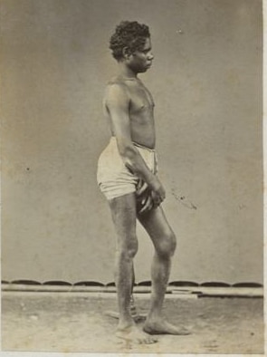 An Aboriginal man standing side on with a his hands clasped in front of him.