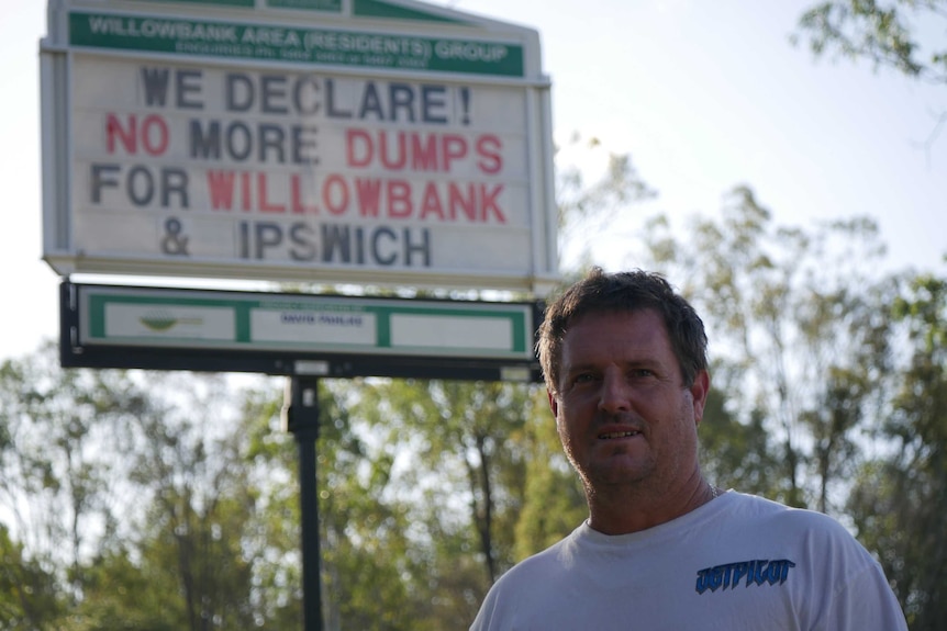 Man in white shirt stands in front of sign that says 'no more dumps for Willowbank and Ipswich'