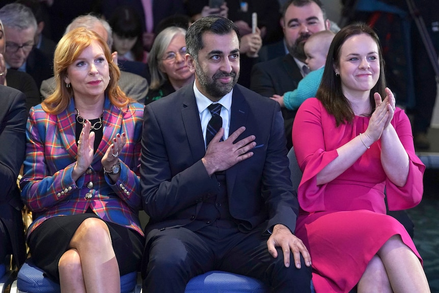 Yousaf hold a hand to his heart as the women on either side of him applaud. 