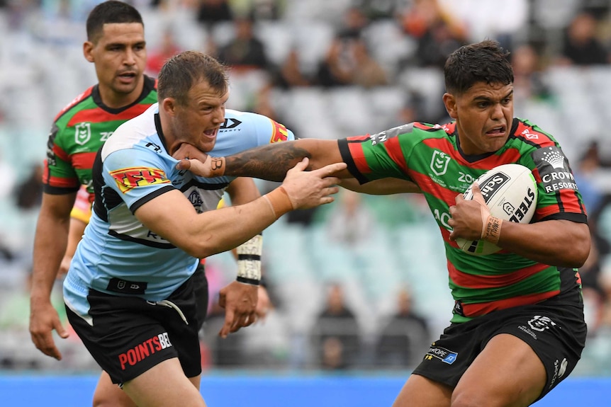 A South Sydney NRL player attempts to pass the ball while holding off a defender.