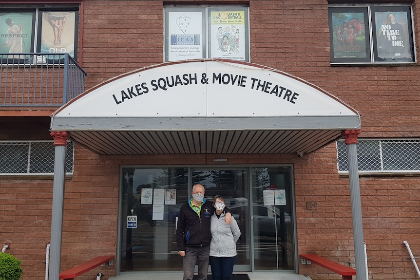 A man and woman wearing masks standing underneath a movie theatre sign.