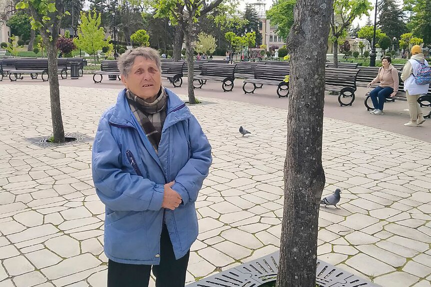 An elderly woman wearing a warm coat stands next to a tree in a paved public space as pigeons walk in the background.