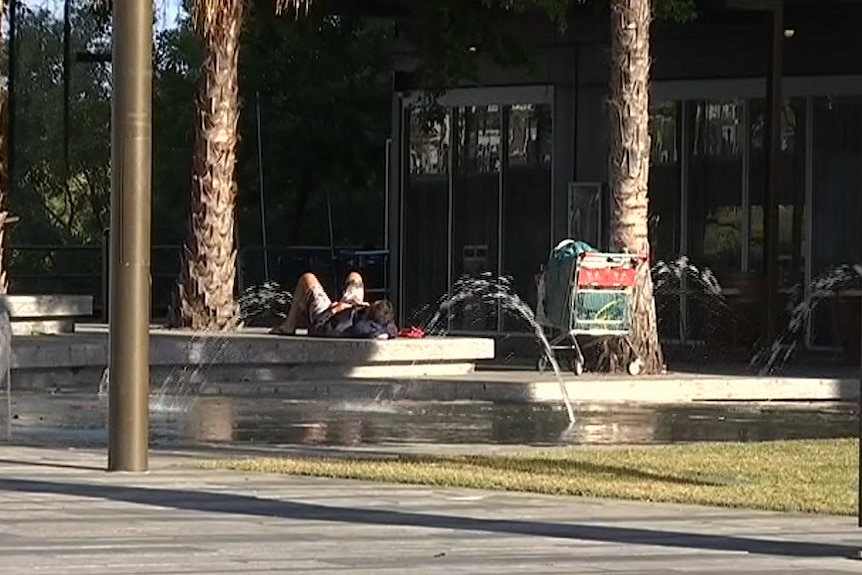 A man lays down on a park bench near a shopping trolley full of possessions.