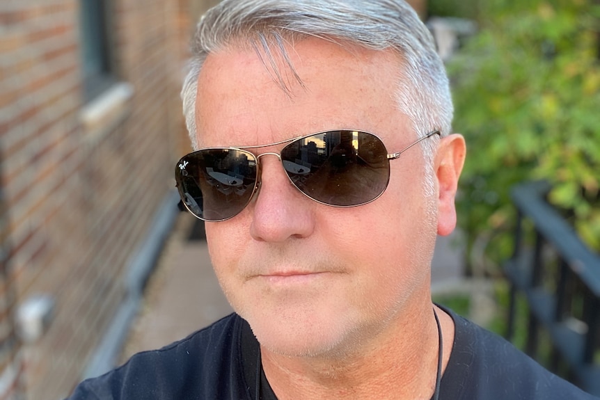 A man with grey hair wearing aviator sunglasses and a navy shirt, taking a selfie.