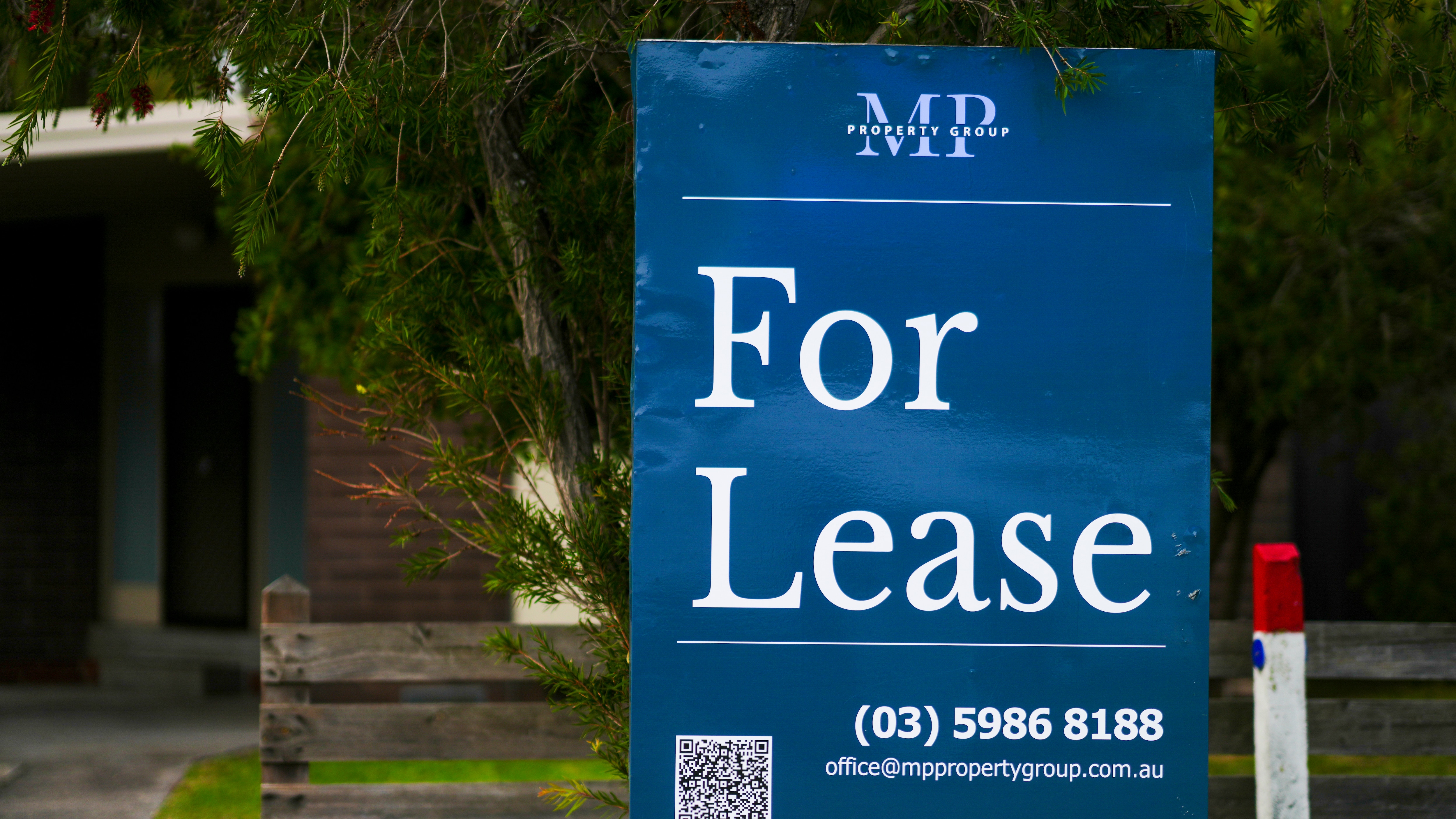 A blue sign with white writing saying "For Lease" and a phone number in front of a tree and wooden fence.