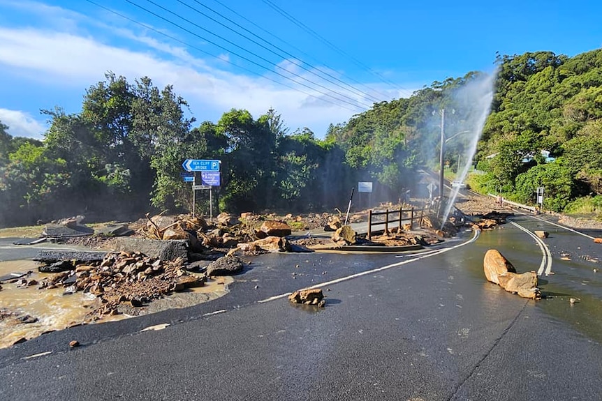 Road damage with rocks, a burst pipe shooting water in the area, rocks strewn across road.