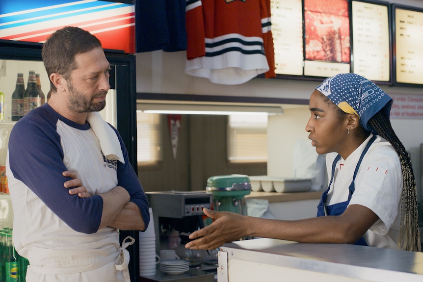 Un hombre blanco con una toalla al hombro y un delantal cruzado de brazos, una joven negra le habla, con la cocina al fondo