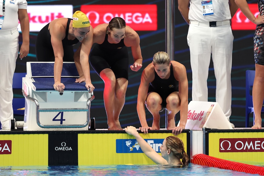 Three female Australian swimmers smile on the blocks and lean down towards their teammate in the water after a race.