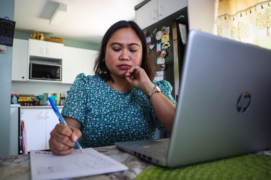 A woman sitting at a table with a computer, writing with a pen on a notebook