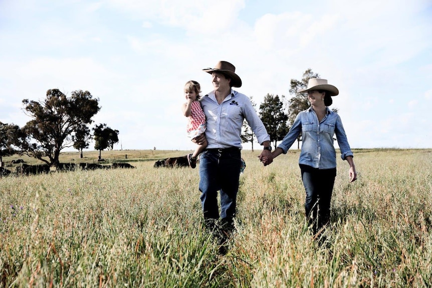 Two farmers, holding hands and a small child, walk through a paddock.
