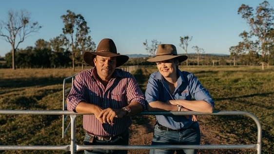 A man in an red and white striped shirt and a woman in a blue shirt lean on a gate with a paddock with trees in the background.