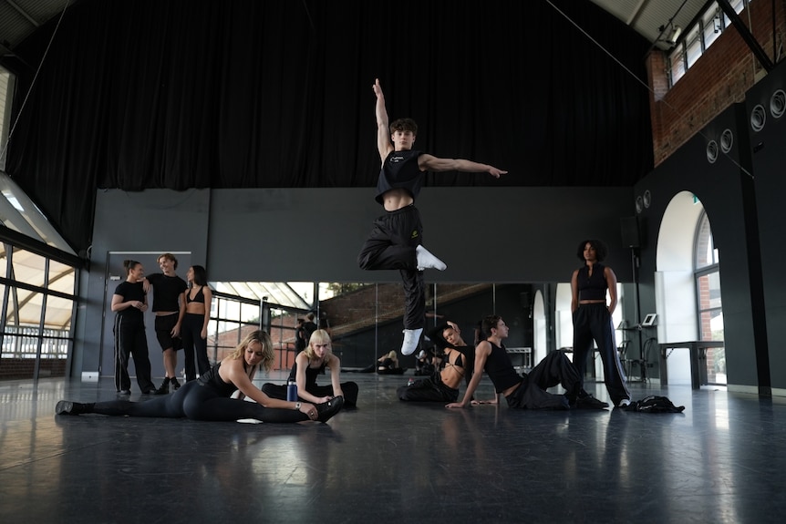 A group of dance students warm up in a large studio, one person is leaping up into the air in the middle